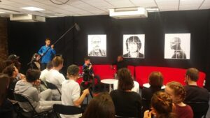 Photographie du plateau de télévision installé pour l'émission avec les portraits des invités et un jeune homme tenant au bout d'une perche le micro