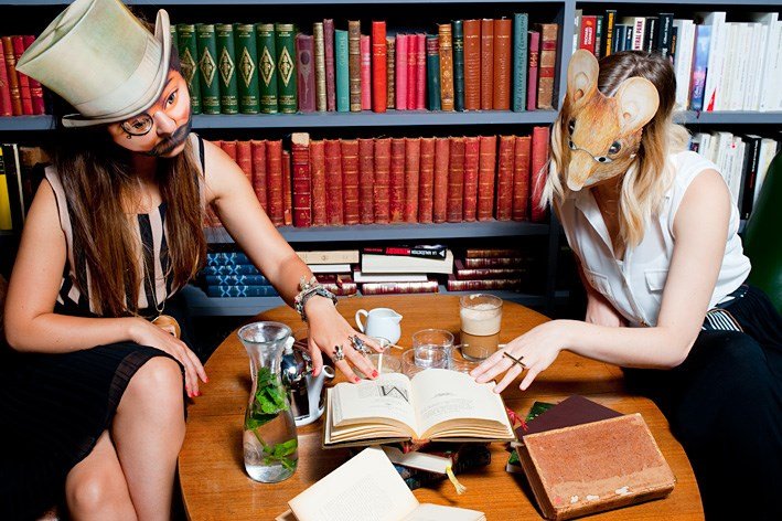 Photographie de deux femmes masquées lisant un même livre devant une bibliothèque